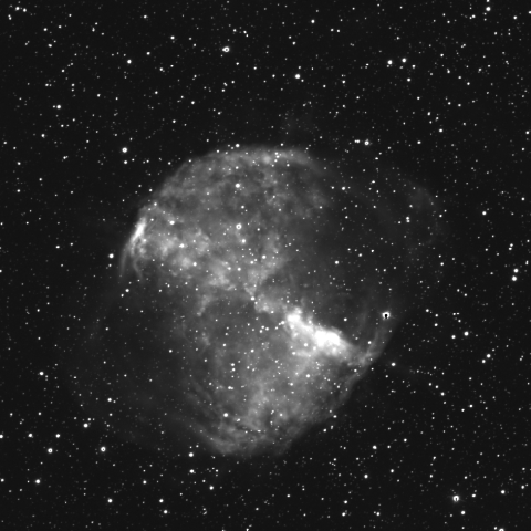 Black-and-white image of a nebula taken through a red filter, showing a roughly circular, cloud-like structure with brighter and darker regions. The nebula is set against a dense background of stars, with intricate textures visible within the nebula's gas and dust