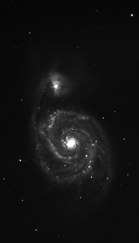 Black-and-white image of the spiral galaxy M51, or Whirlpool Galaxy, taken through a blue filter. The galaxy features a bright central core with well-defined spiral arms extending outward, containing regions of varying brightness. A smaller companion galaxy appears near the top, connected by a faint bridge of material