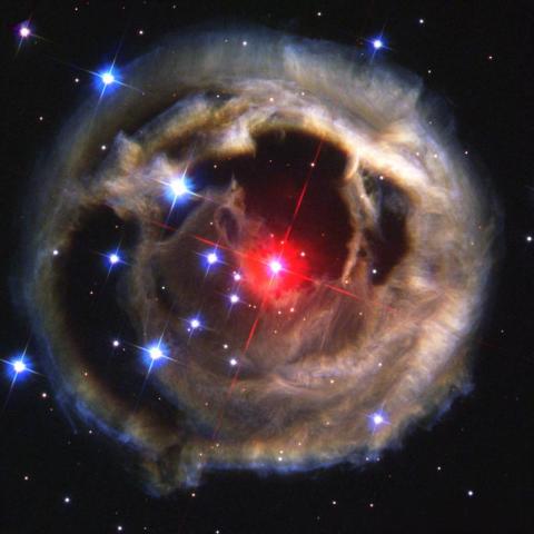 An image of a glowing red star surrounded by a dusty, circular nebula with bright blue stars scattered throughout the dark space background