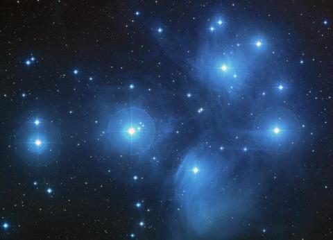 An image of the Pleiades star cluster, showing a collection of bright blue stars surrounded by a faint blue nebula in the vast dark expanse of space