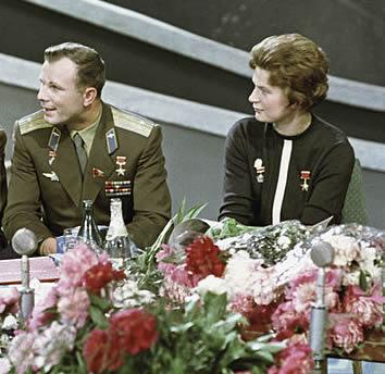 A color photograph of Yuri Gagarin and Valentina Tereshkova sitting at a table adorned with flowers. Gagarin, in military uniform, and Tereshkova, dressed in a dark outfit with a medal, are both Soviet space pioneers. They appear to be at a formal event, possibly celebrating their achievements in space exploration