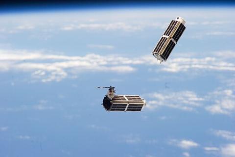 Two CubeSats, small rectangular satellites, floating in space above the Earth's atmosphere with clouds visible in the background