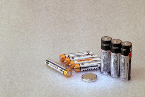 A few various sized batteries laying on a surface, with three stood upright. There is also a small, circular, silver cell as well.