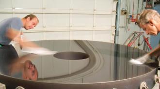 Two technicians polishing a 2m telescope mirror, carefully cleaning its reflective surface in a workshop. The reflection of one technician is visible on the mirror as they work 