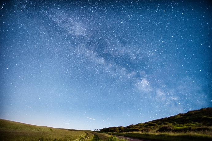 This image shows a clear night sky with a vast number of stars visible, including a distinct view of the Milky Way stretching across the horizon. The foreground features an open field and some hilly terrain