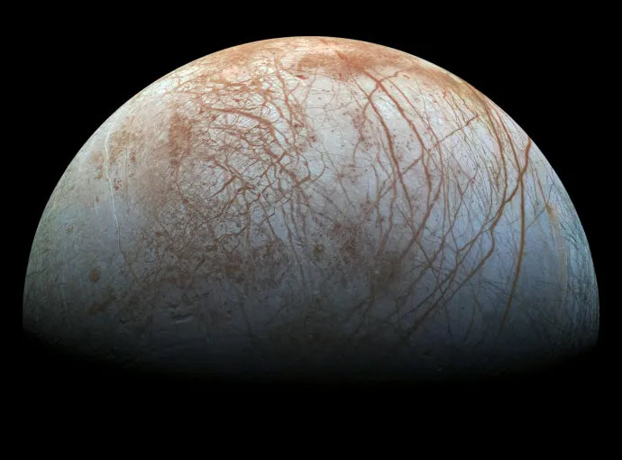 The image shows Europa, one of Jupiter's moons. It appears as a round celestial body with a smooth, icy surface. The surface is marked with reddish-brown lines and patches, which are likely cracks or ridges in the ice. The background is black, emphasizing the moon's pale, textured appearance.