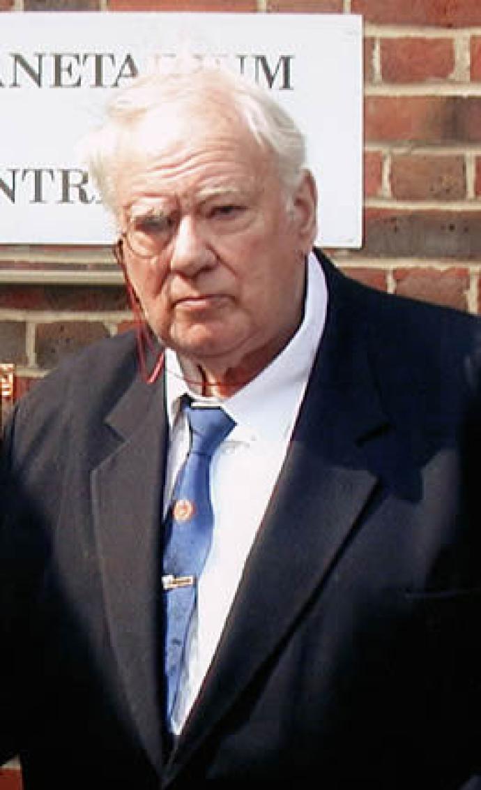 A photograph of Sir Patrick Moore, the British astronomer and broadcaster, wearing a dark suit and blue tie. He appears serious, standing in front of a brick building with part of a sign visible in the background