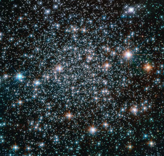 A dense star cluster filled with thousands of bright stars, some shining with a blueish hue, while others emit a warmer orange glow, set against the blackness of space.