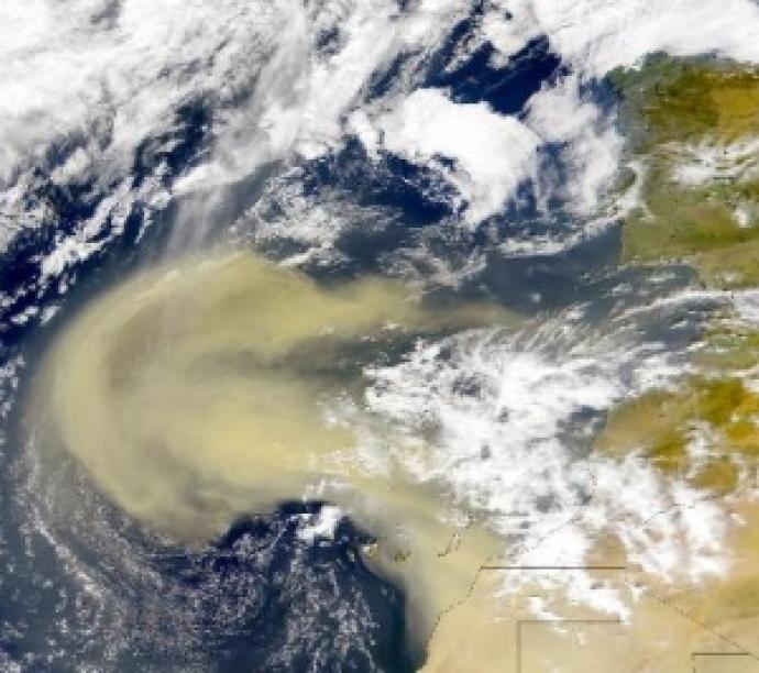 A satellite image shows a large, yellowish dust cloud spreading over the ocean from the African coast