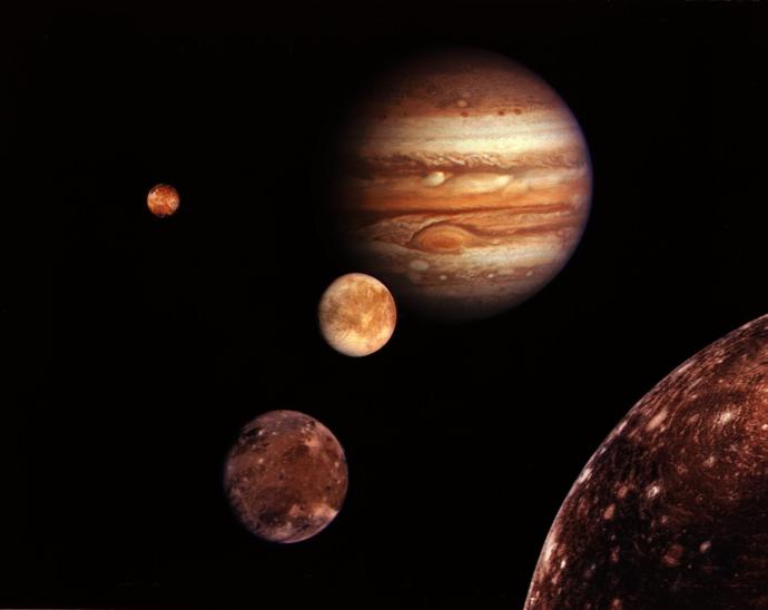 Image of Jupiter with its four largest moons visible in the foreground. The planet displays its distinct bands and Great Red Spot. The moons, from left to right, are Io, Europa, Ganymede, and Callisto